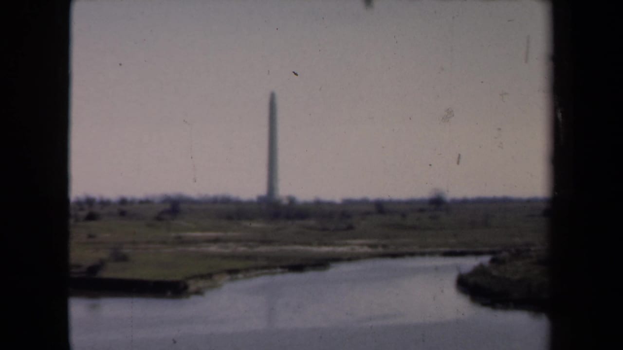1946 8mm frame showing a distant vertical monument near a shoreline, viewed across a river or canal through dark foreground edges.