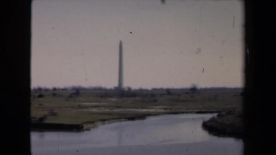 1946 8mm frame showing a distant vertical monument near a shoreline, viewed across a river or canal through dark foreground edges.