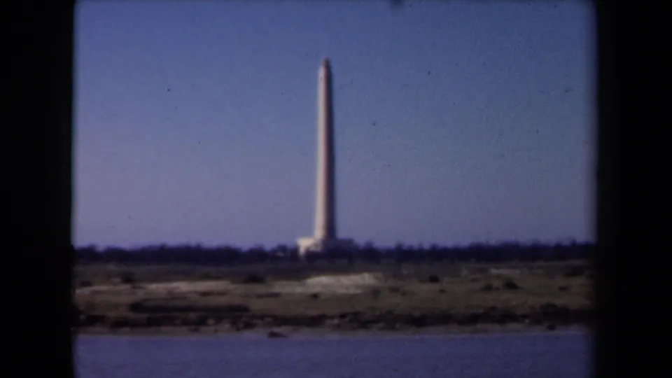 1946 Netherlands 8mm frame showing a distant tall tower or beacon rising above low scrubland across a calm body of water.