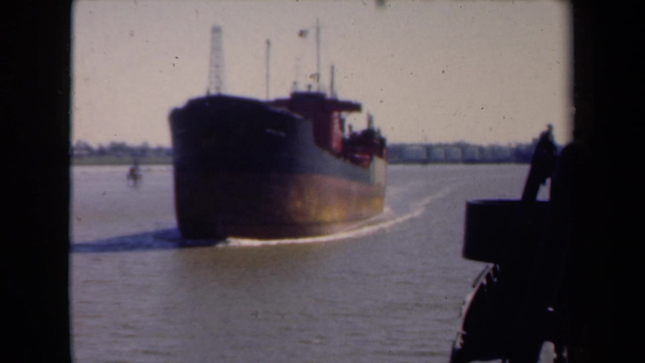Large dark cargo ship approaching on a river with wake and distant shoreline.