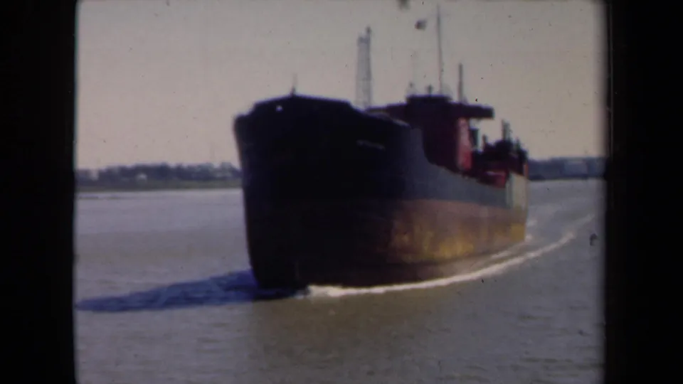 A 1946 Netherlands 8mm frame shows the cargo vessel passing close by, with river wake, pale sky, and distant industrial shoreline.
