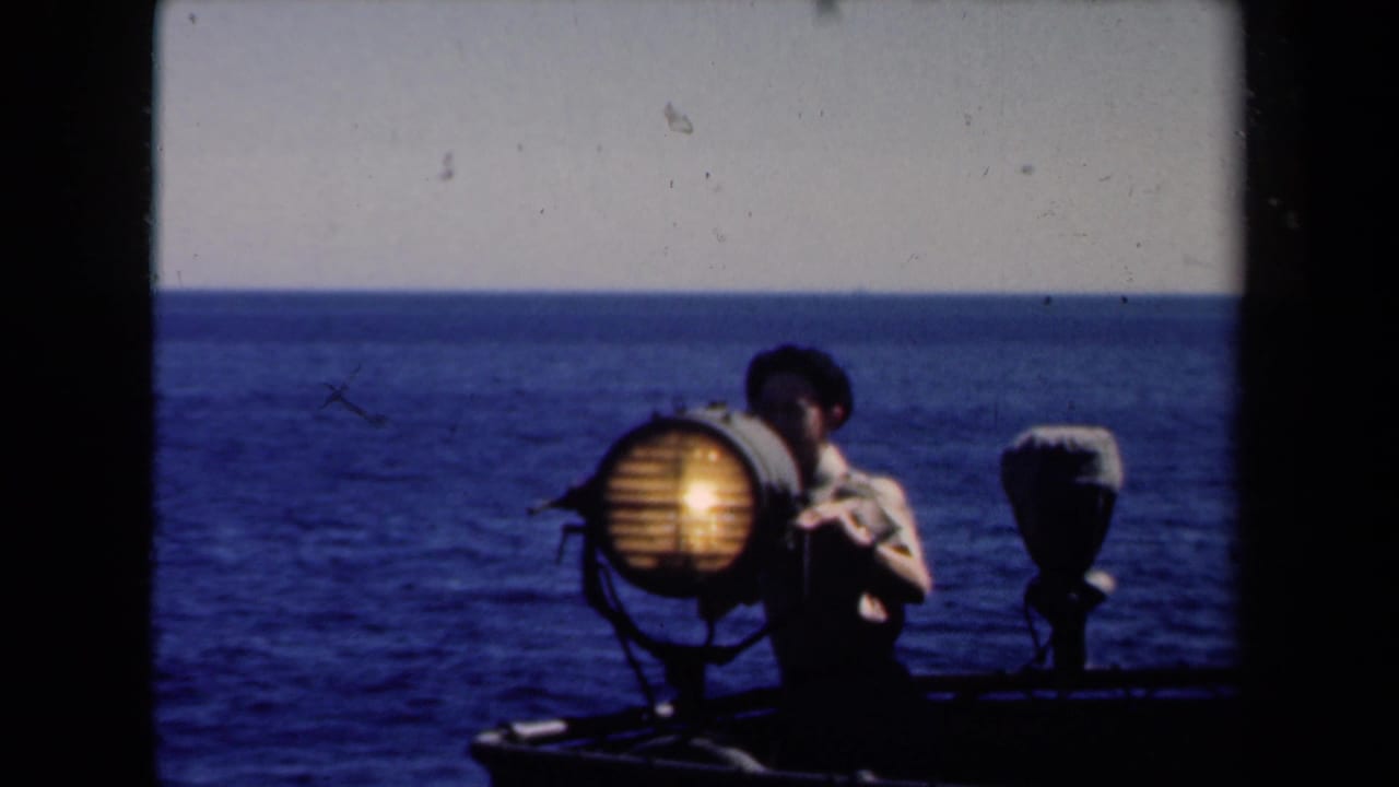 1946 Netherlands maritime frame showing a sailor operating or standing beside a lit searchlight against an open sea horizon.