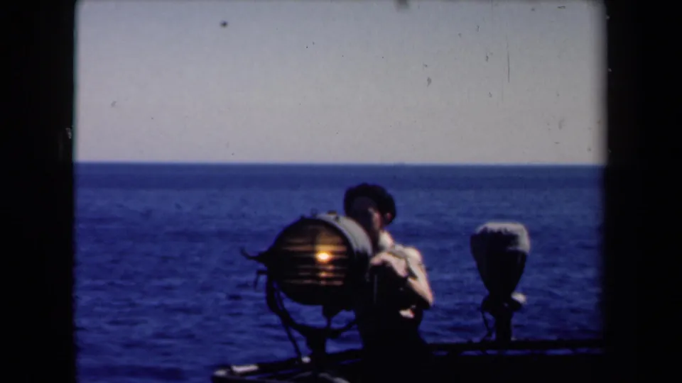 In Netherlands 1946 home-movie footage, a young man stands near a glowing round searchlight on a ship deck facing the open sea.