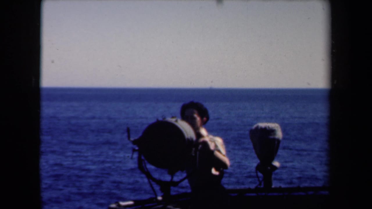 Young man on deck with a dark searchlight shape and ocean horizon behind him.