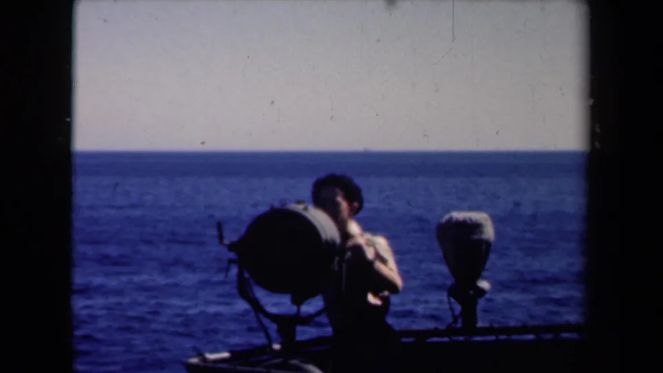 A large shipboard searchlight fills the foreground while the young man stands partly obscured, with open water stretching behind him.