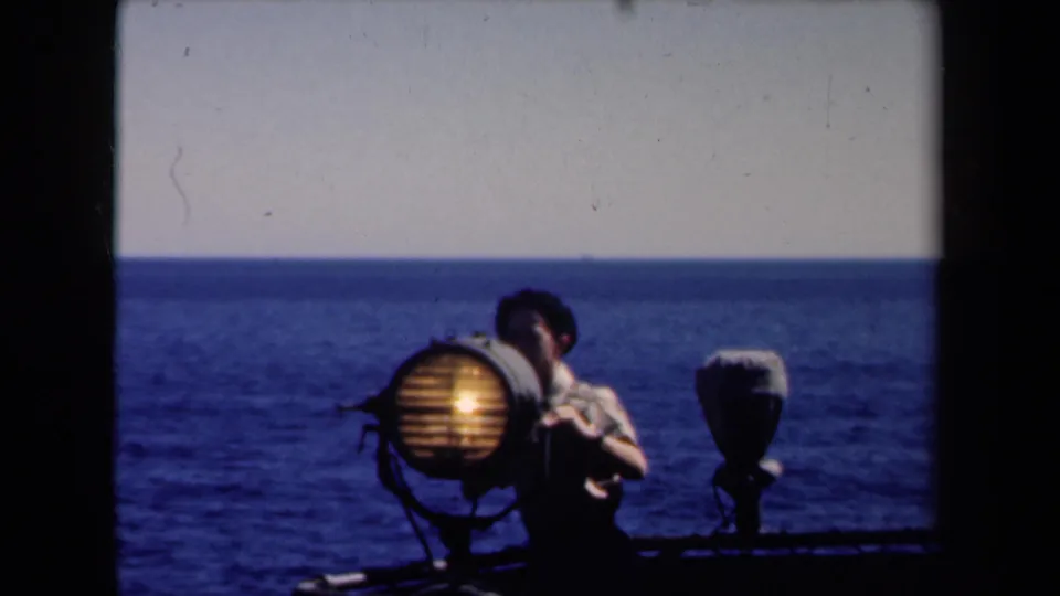 A glowing round searchlight sits beside the young man on the deck, with a second fixture, railing, and ocean horizon visible behind.
