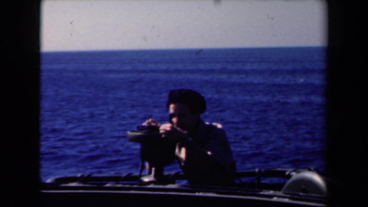 Man in dark beret leans toward a mounted optical device on a boat with open sea behind him.