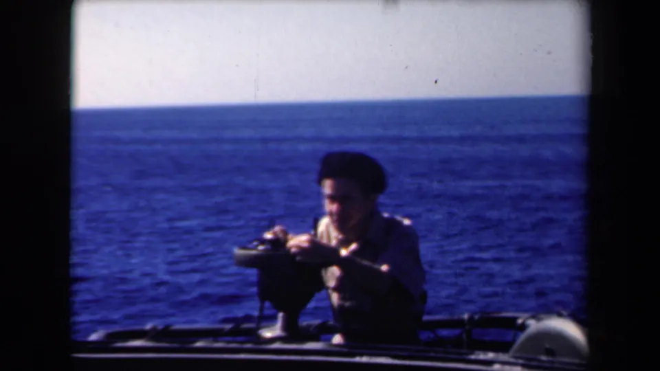The 8mm frame shows the man at the mounted instrument, framed by boat rails, pale sky, and open water.