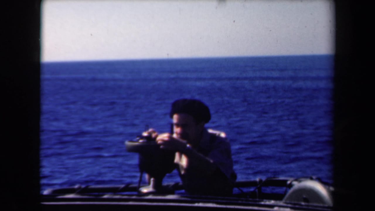 Sailor in a dark cap leaning over a mounted gun with open ocean behind him.