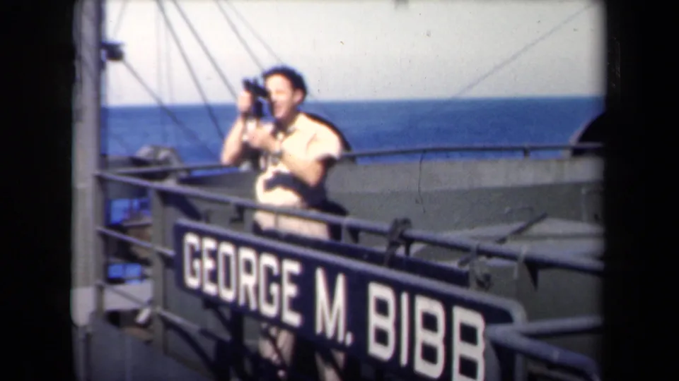 A man on the George M. Bibb deck raises a camera or optical device, with rigging, railings, and sea visible behind him.