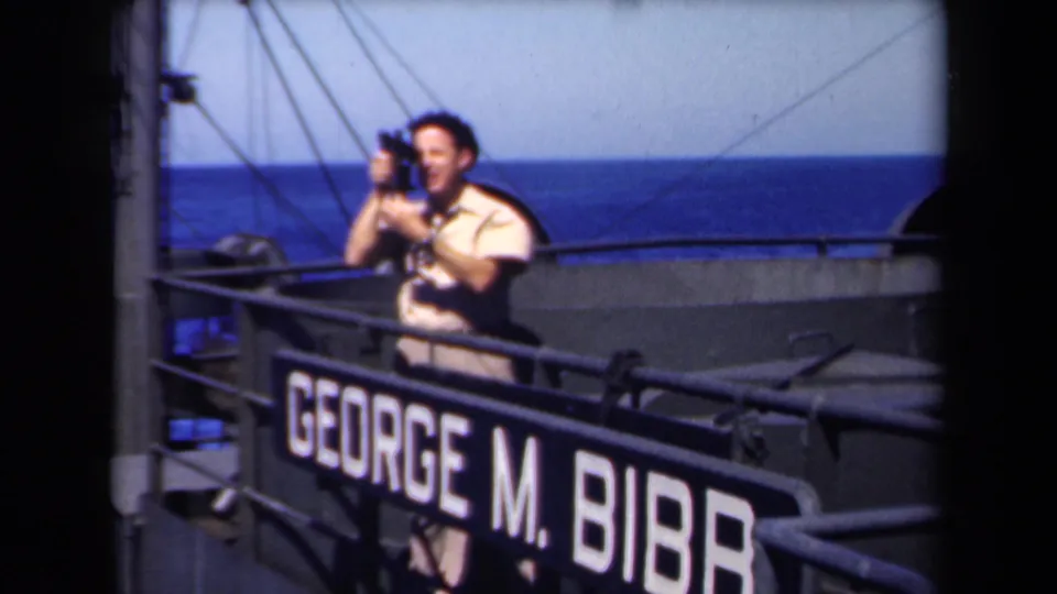 1946 shipboard frame showing a man filming or looking through a camera beside the George M. Bibb rail sign and ocean horizon.