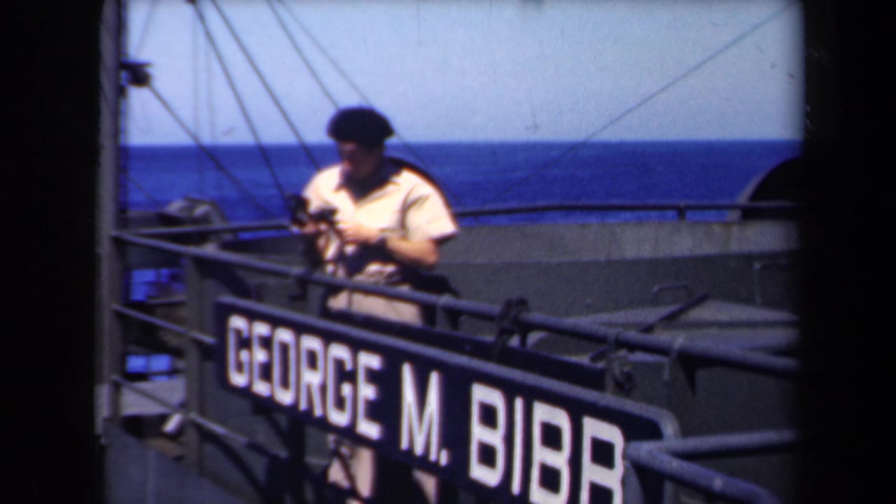 Seaman in a dark beret looks down while adjusting a camera on the ship deck.