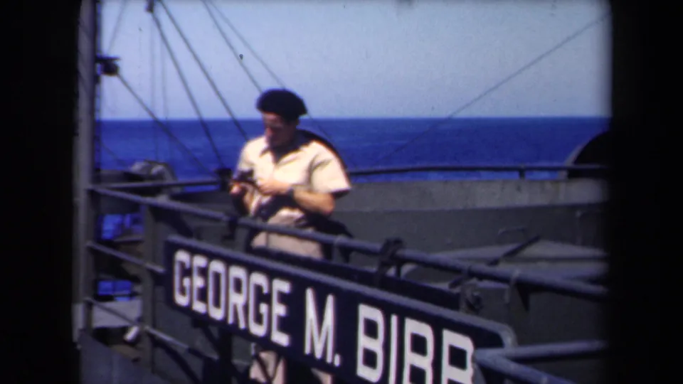 The 1946 8mm frame shows a seaman handling a camera near the George M. Bibb nameplate, with ocean visible beyond the rail.