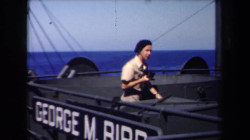 A young seaman carries a camera along the ship deck in 1946 home-movie footage, with open sea behind the George M. Bibb nameplate.