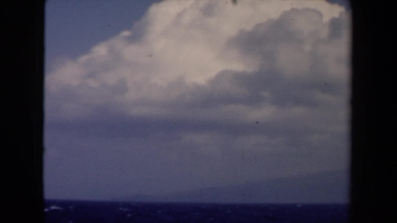 Blue ocean horizon with distant land below large white clouds, framed by dark 8mm film edges.