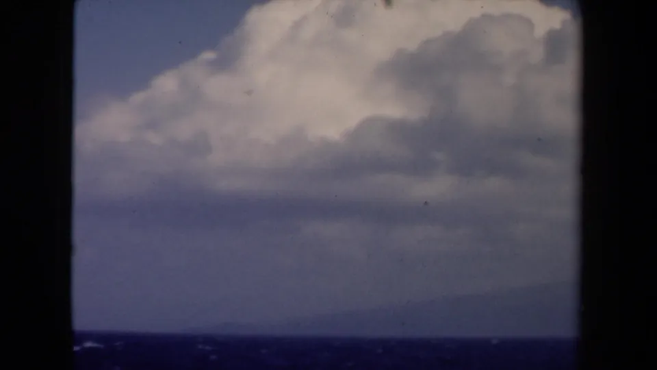 1946 Netherlands 8mm frame showing a blue sea horizon, faint distant land, and large cumulus clouds from a shipboard viewpoint.