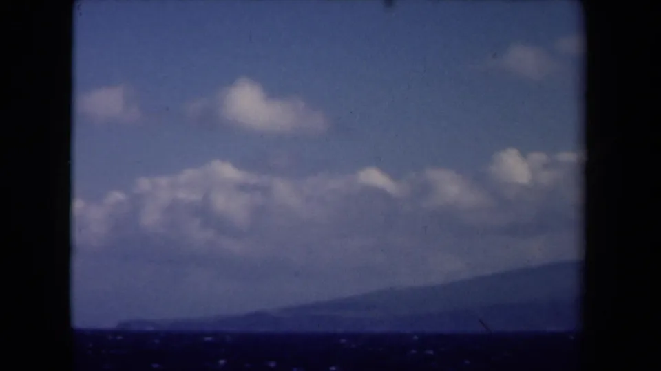 A 1946 8mm frame from the Netherlands showing blue sky, scattered clouds, and a distant sloping landform beneath dark window framing.