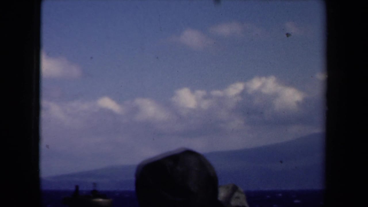 Silhouetted passenger hat at the lower frame edge with ocean, distant coastline, clouds, and blue sky.