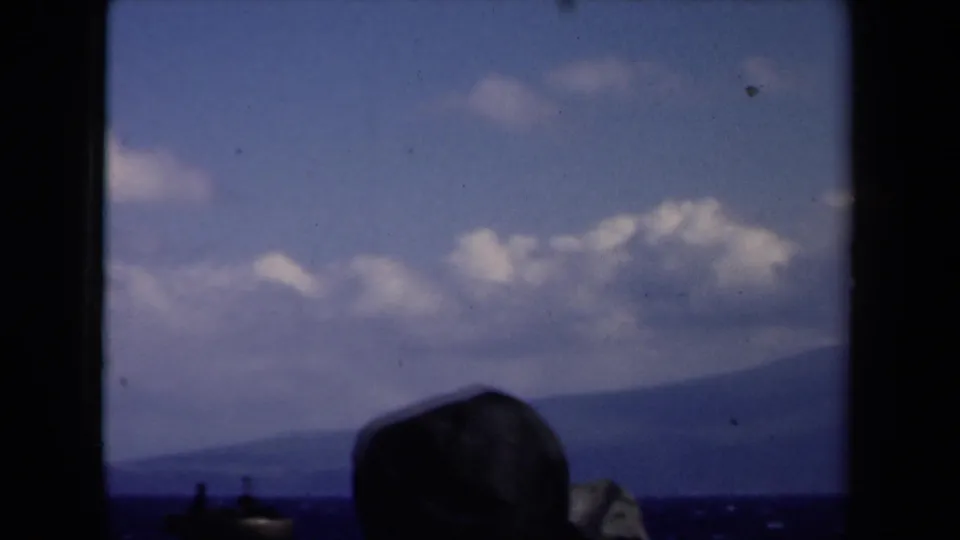 1946 8mm Netherlands home-movie frame showing a silhouetted passenger hat against open water, distant mountainous coastline, clouds, and blue sky.