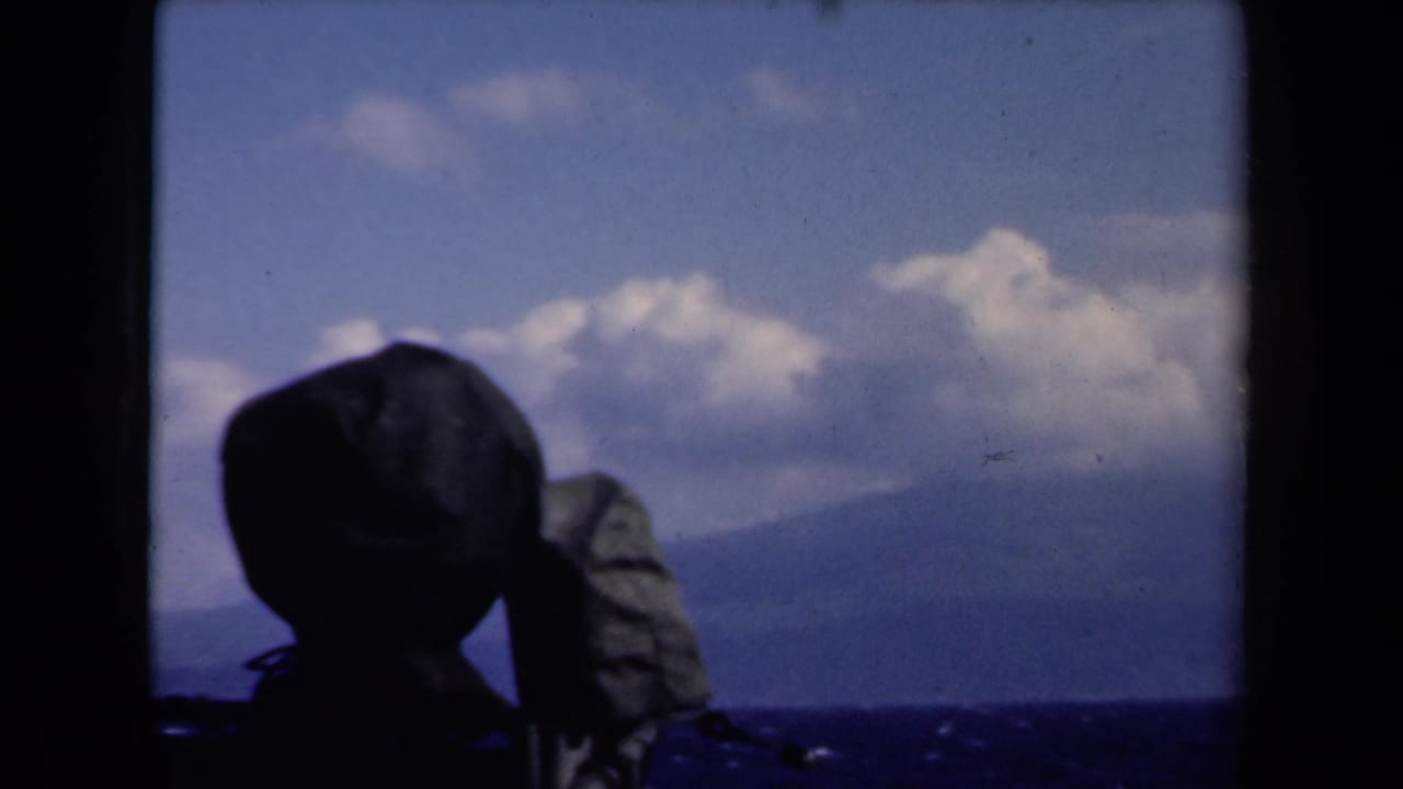 Dark hat silhouette and shoulder near the horizon with open sea, distant landforms, clouds, and sky.