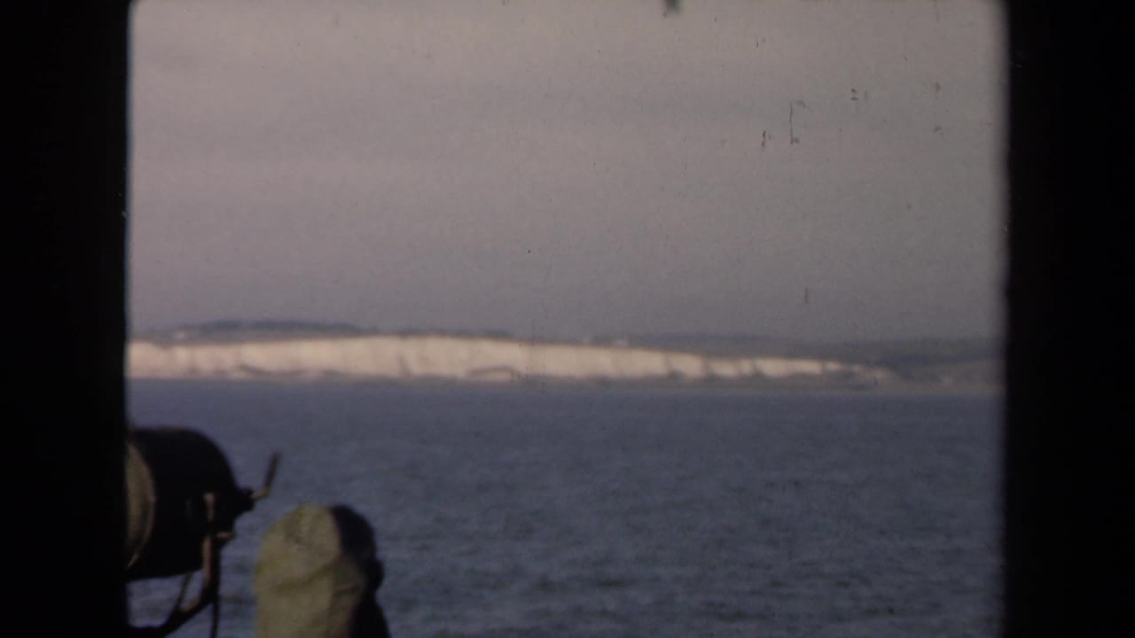 1946 8mm view from a ship window toward pale coastal cliffs across calm water, with a small deck fixture at lower left.
