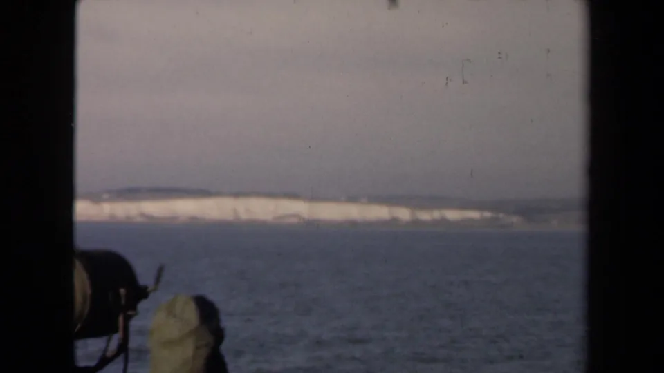 1946 8mm view from a ship window toward pale coastal cliffs across calm water, with a small deck fixture at lower left.