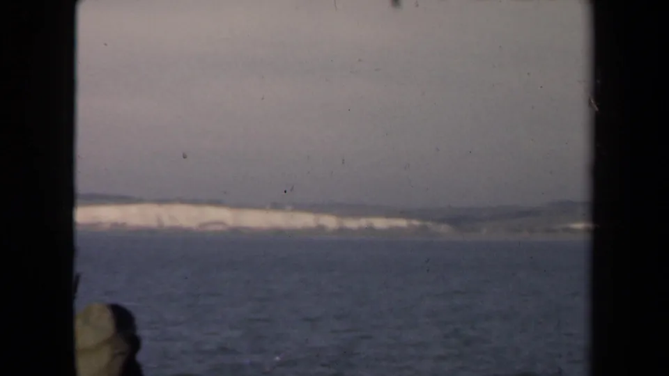Distant white cliffs and low hills appear across calm water, viewed through the dark framing of a ship window or porthole.