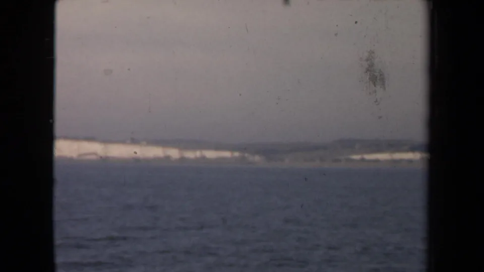 A shipboard 8mm frame shows calm water, a pale cliff line, and dark window edges, with visible dust and faded color in the scan.