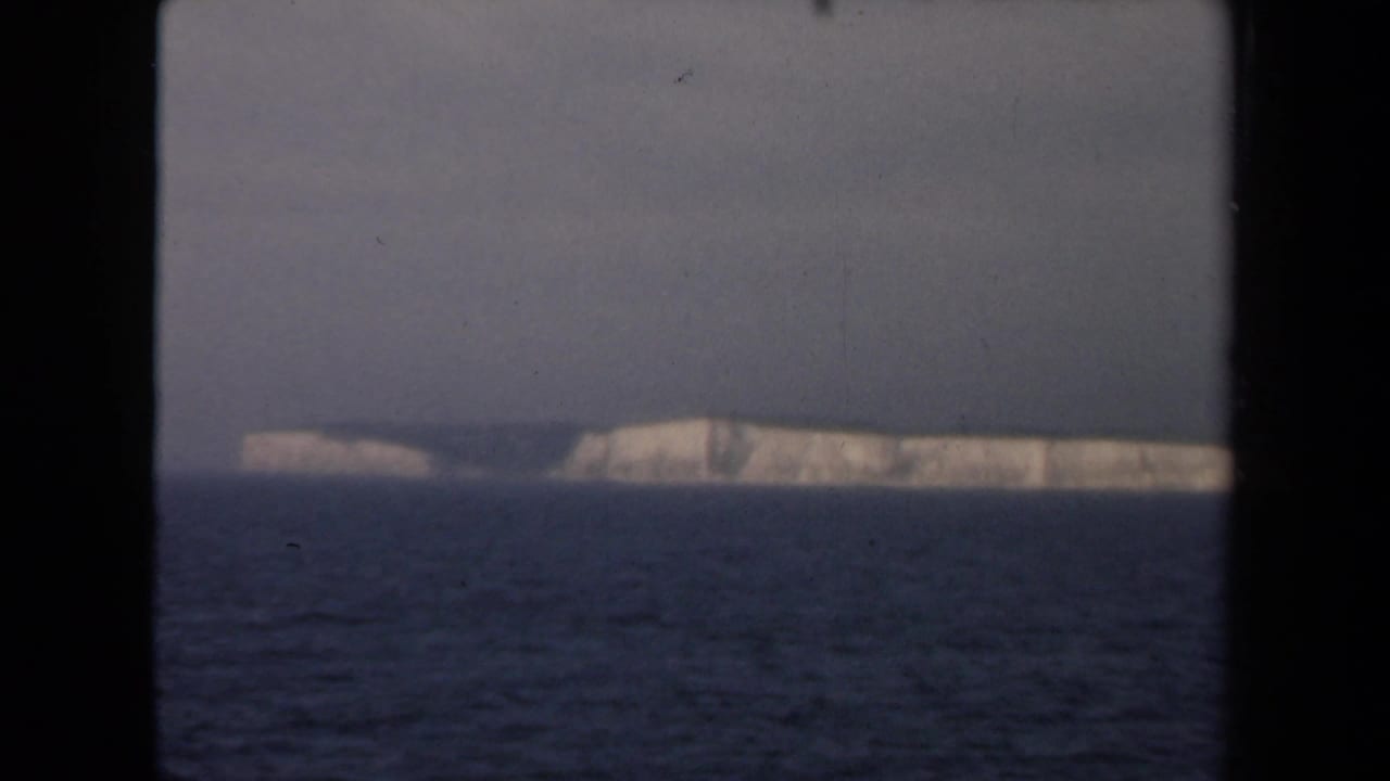 Distant white cliffs across dark water under a gray sky, framed by black borders.