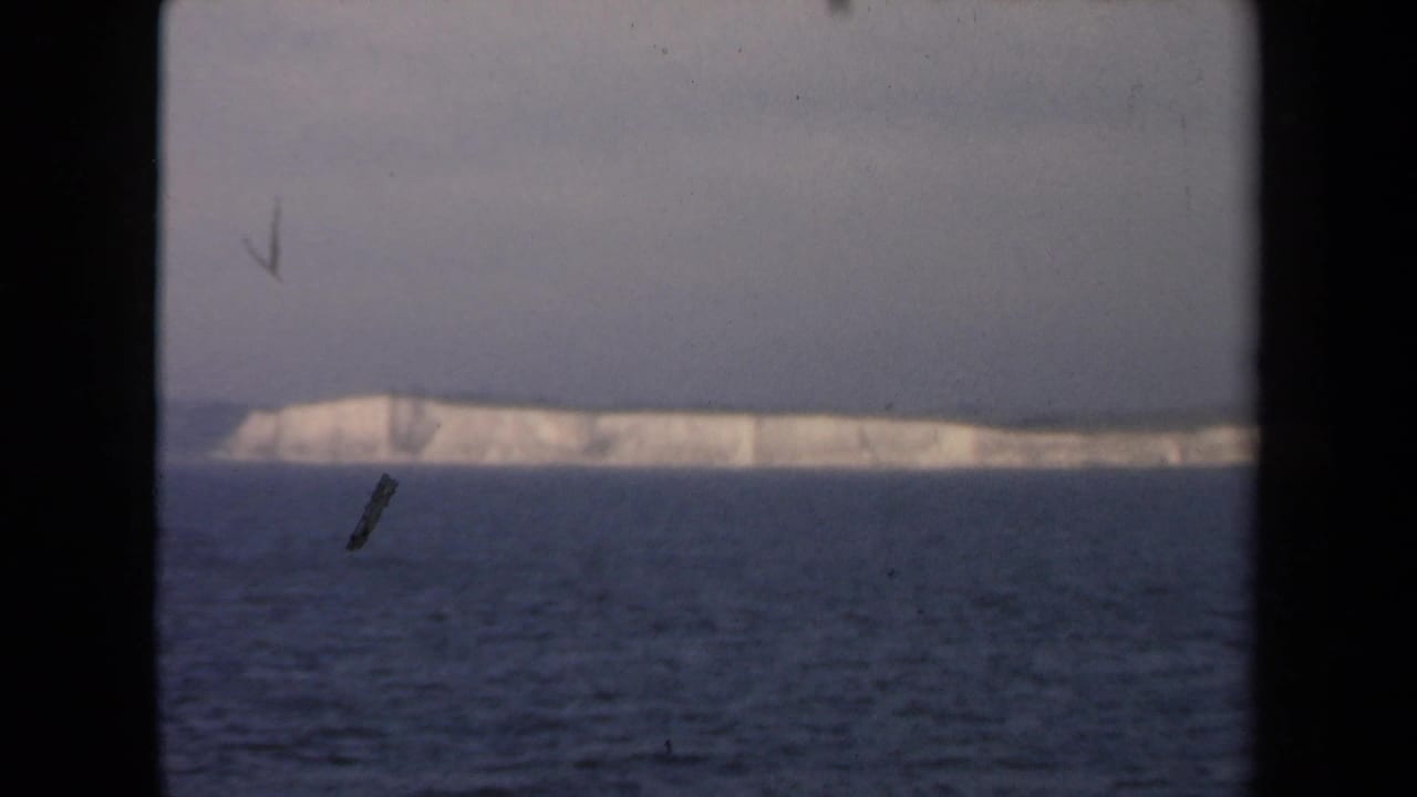 Distant pale ice mass across dark ocean under a gray sky, framed by black film borders.
