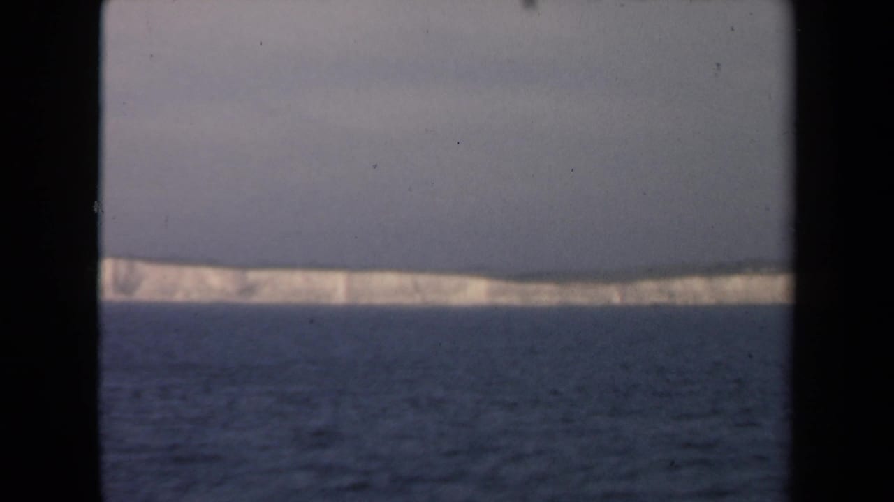 Archival 8mm view of a long pale iceberg or ice edge on the horizon, seen across textured ocean water with a dark vertical border.