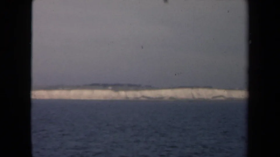 A muted 8mm frame holds on a low white ice-like coastline beyond dark water, with black gate edges and visible film wear.