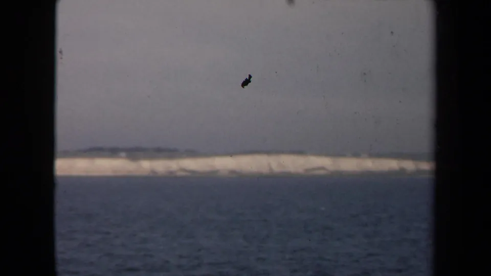 Final 8mm frame of a distant pale ice or coastal formation across water, with visible dust, grain, and black frame borders.