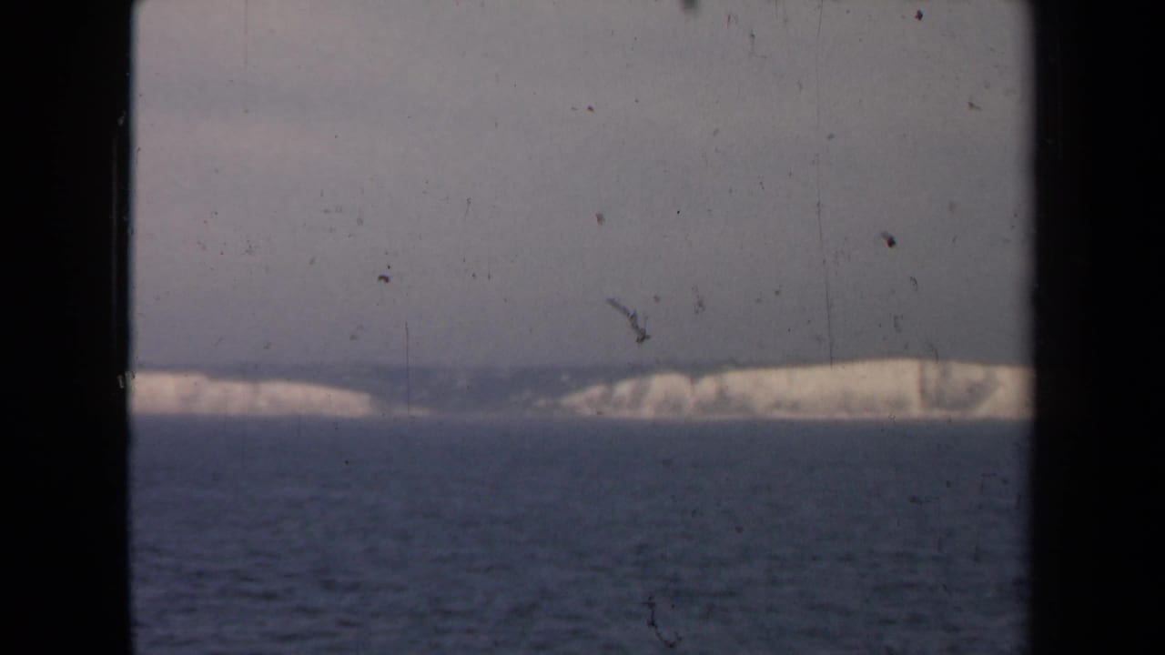 Open water and a low pale coastline seen through dark vertical framing with film scratches.