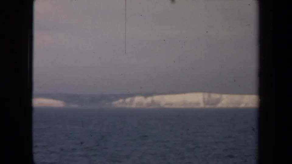 1940s 8mm frame with a distant pale landmass across blue water, shot through a dark window-like edge.