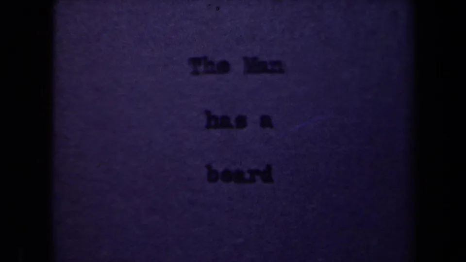 1940s 8mm frame showing a dim typed title card reading The Man has a beard, with heavy grain and dark vertical film borders.