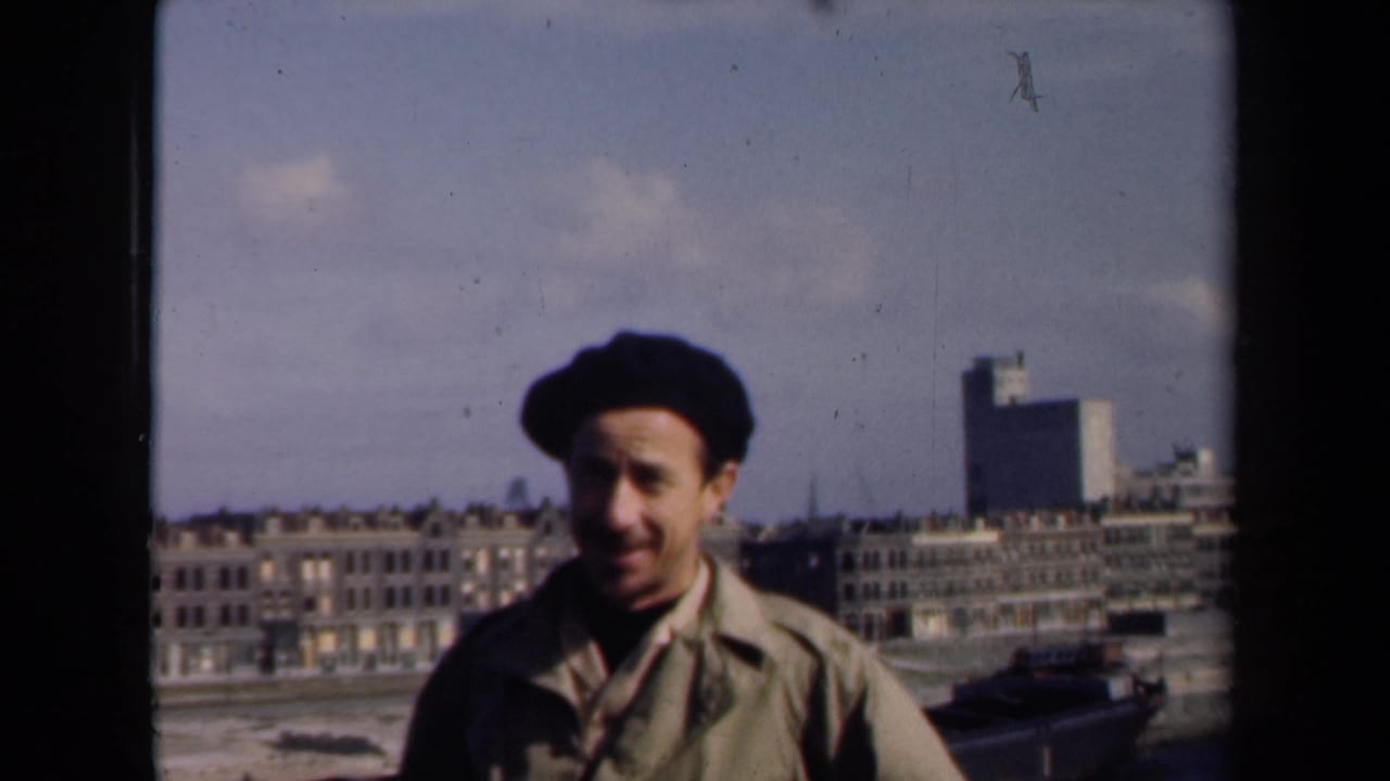 Smiling man in beret stands in front of rows of city buildings.