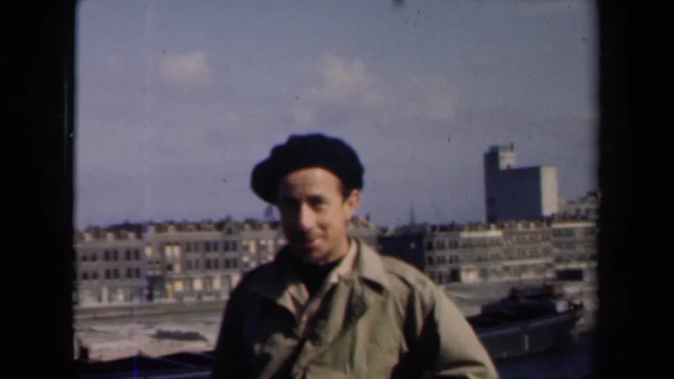 A man in a dark beret is filmed close to camera with a postwar Netherlands cityscape visible behind him.