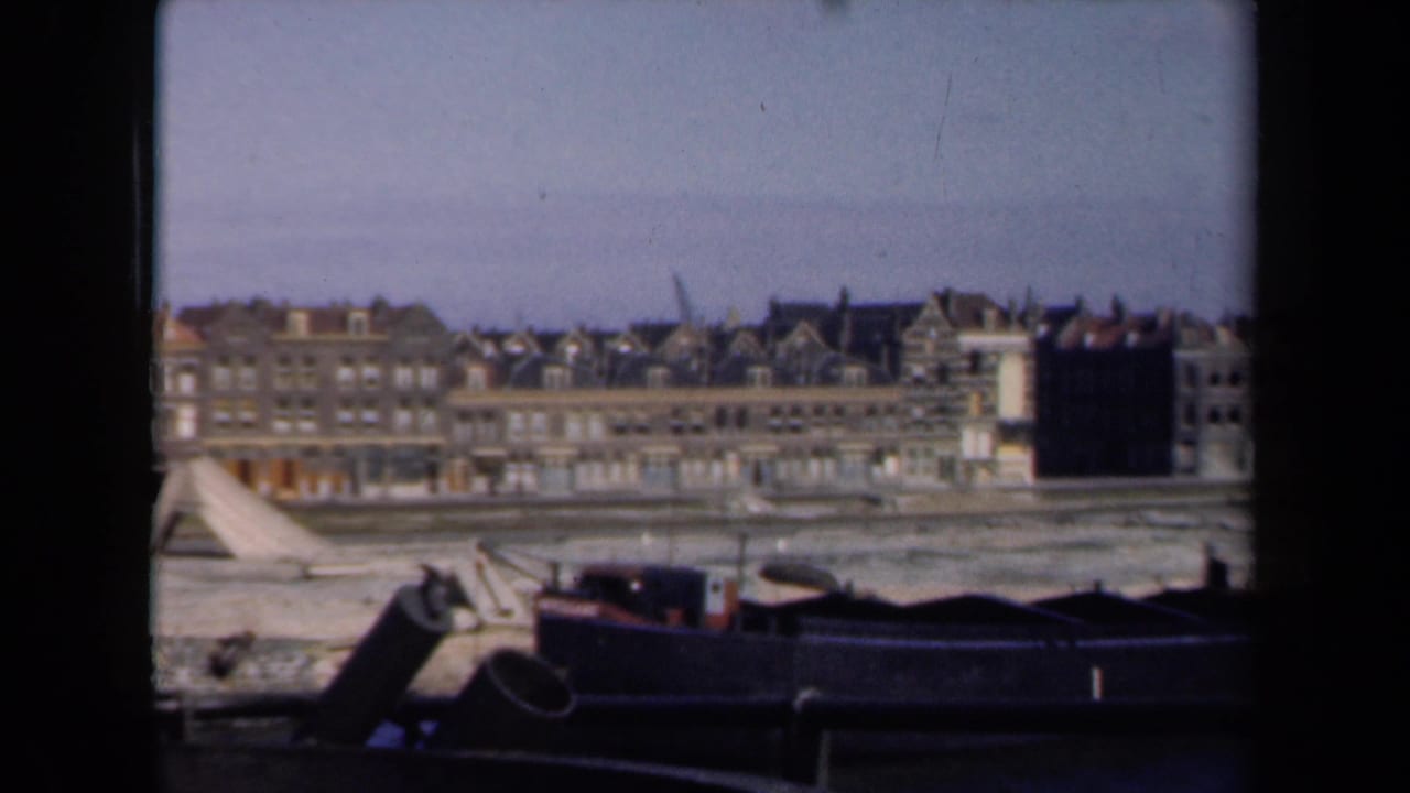 1946 Netherlands home-movie view of waterfront buildings, gabled roofs, muddy shore, and dark barges in the foreground.