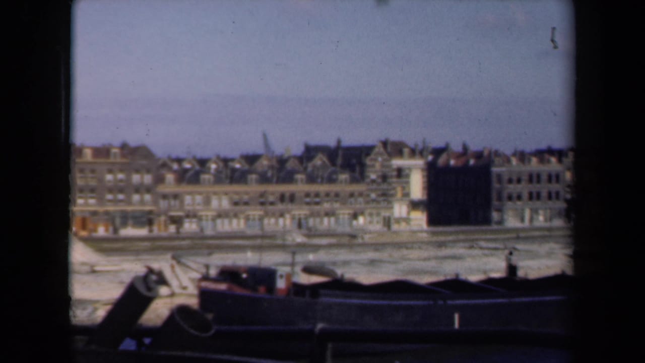 Row of waterfront buildings behind dark barges, with a small post or figure-like shape at right.