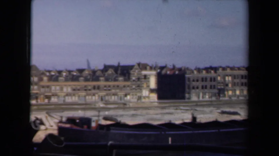 Soft 8mm view of a Dutch waterfront block in 1946, with gabled rooftops, pale facades, muddy shore, and dark boats below.