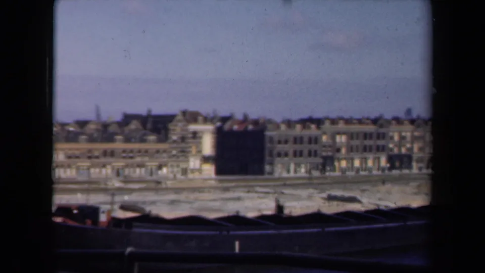 1946 Netherlands waterfront cityscape with a row of buildings, gabled roofs, dark barges, and clear sky in muted 8mm color.