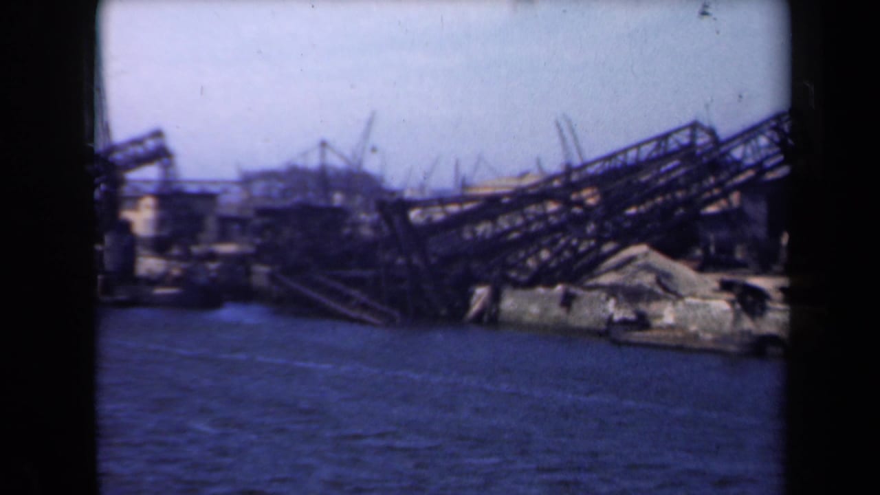 Twisted waterfront framework and rubble seen across blue water under a pale sky.