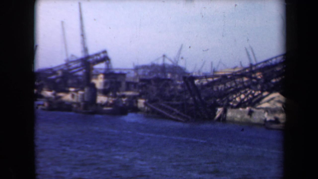 1946 Netherlands 8mm view across water toward a shipyard with cranes, angled metal framework, low buildings, and a towered structure.