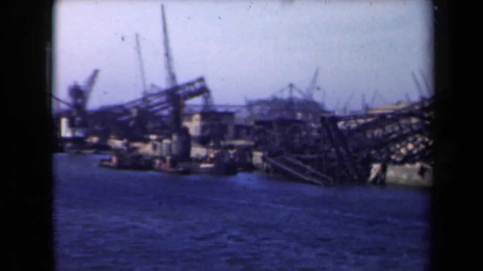 1946 Netherlands waterfront view showing dock cranes, low industrial buildings, dark angled framework, and calm blue water in the foreground.
