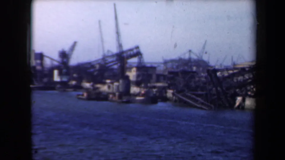 Soft 8mm frame of a Dutch shipyard in 1946, with harbor water, cranes, a towered gray building, and tilted metal framework.