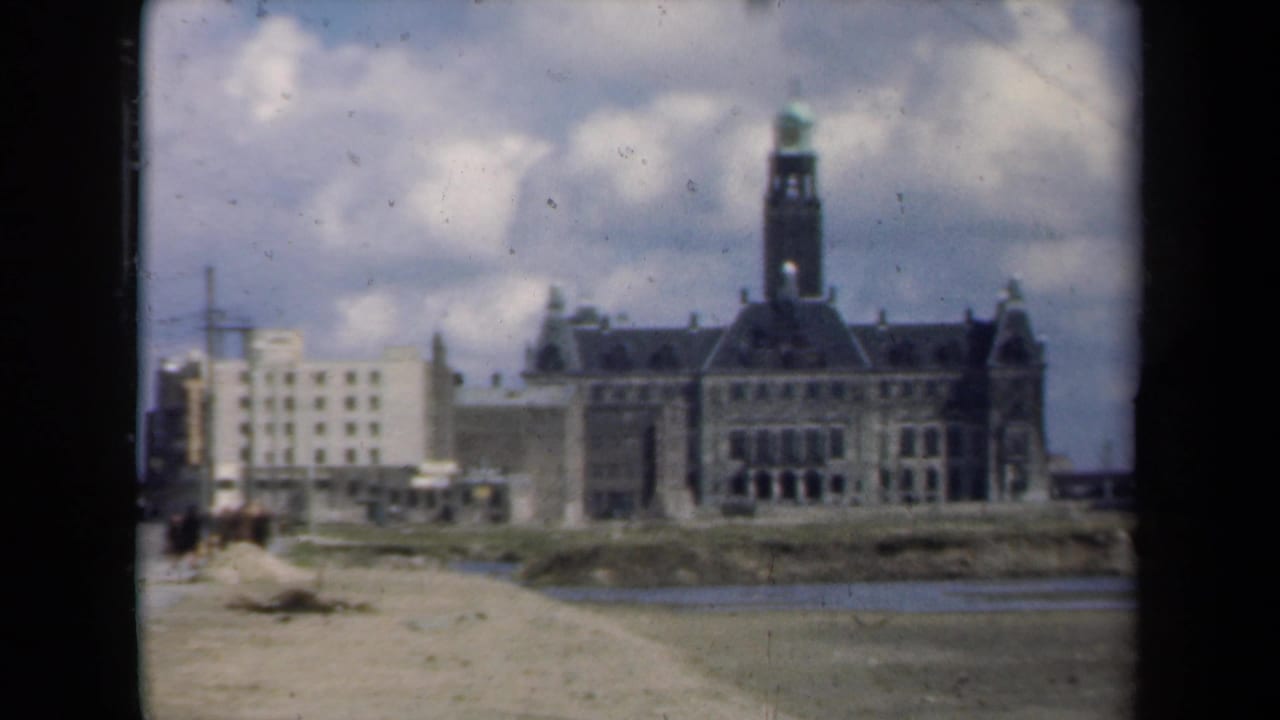 1946 Netherlands home-movie view of a large towered building across a calm river, with a pale modern building at left and cloudy sky above.