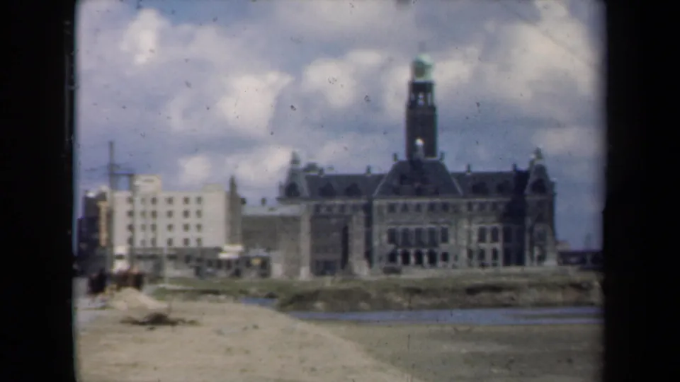 1946 Netherlands home-movie view of a large towered building across a calm river, with a pale modern building at left and cloudy sky above.