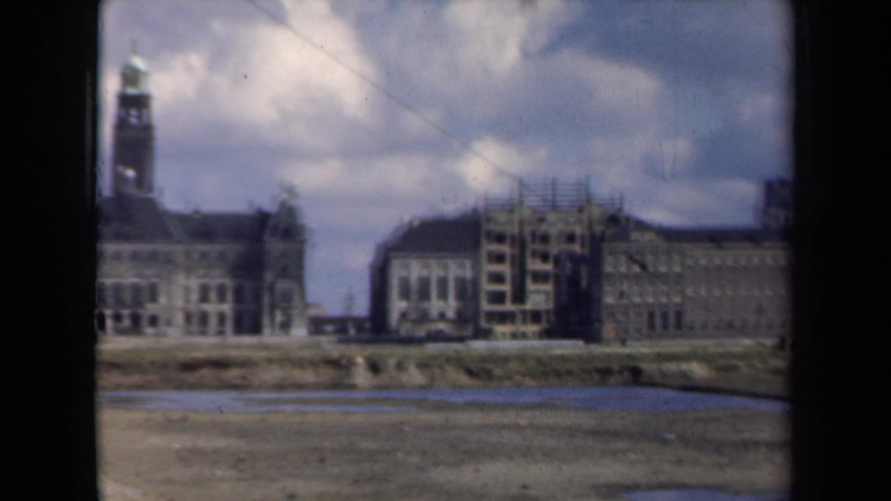 1946 Netherlands 8mm frame showing distant civic buildings, a scaffolded facade, a tall tower, cloudy sky, and muddy foreground.