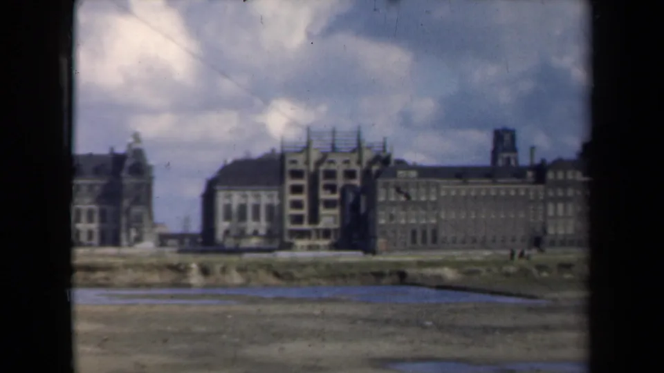 1946 Netherlands frame with a broad distant building frontage, central scaffolding, tower silhouettes, open muddy ground, and soft 8mm color.